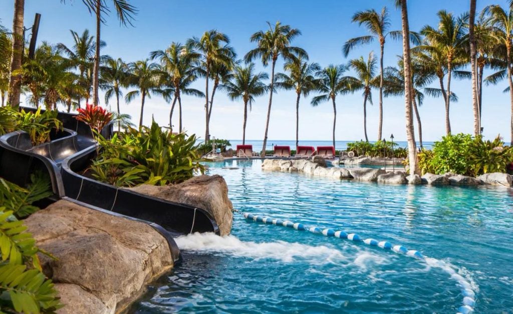 The Helumoa Playground Pool at the Sheraton Waikīkī, available to Royal Hawaiian guests, featuring a winding water slide, tropical landscaping, and a large lagoon-style pool lined with palm trees and red lounge chairs overlooking the ocean.