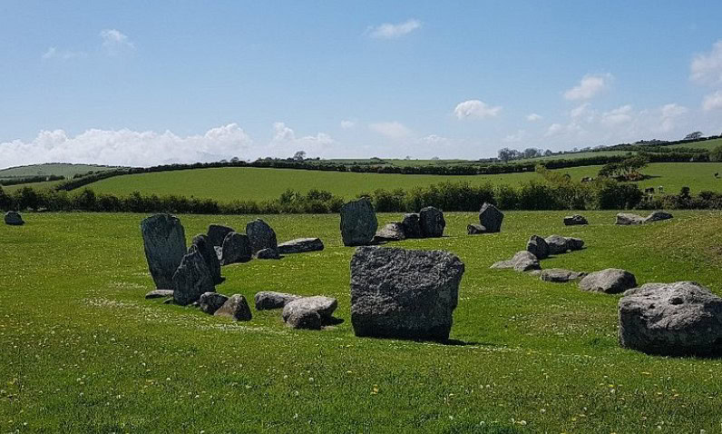 The Ballynoe Stone Circle in Ireland, an ancient megalithic site, is set in a lush green field surrounded by rolling hills and hedgerows. The large, weathered stones form a circular arrangement, standing and lying in various positions. The bright blue sky with scattered clouds enhances the peaceful and mystical atmosphere of this historic landscape.