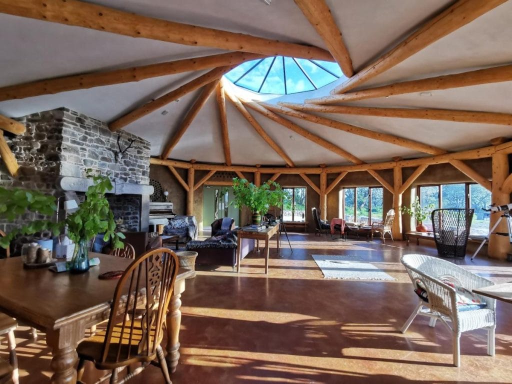 The interior of Lough Mardal Lodge in Donegal, Ireland, featuring a unique circular design with exposed wooden beams radiating from a central skylight. The cozy space includes a stone fireplace, rustic wooden furniture, and large windows allowing natural light to flood in. Lush greenery and inviting seating areas enhance the eco-friendly retreat's warm atmosphere.