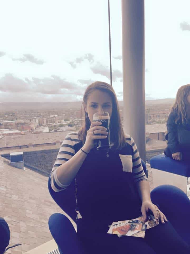 Phoenyx sipping a pint of Guinness while seated by a large window at the Gravity Bar in the Guinness Storehouse, Dublin, Ireland, with panoramic views of the city and cloudy skies in the background.