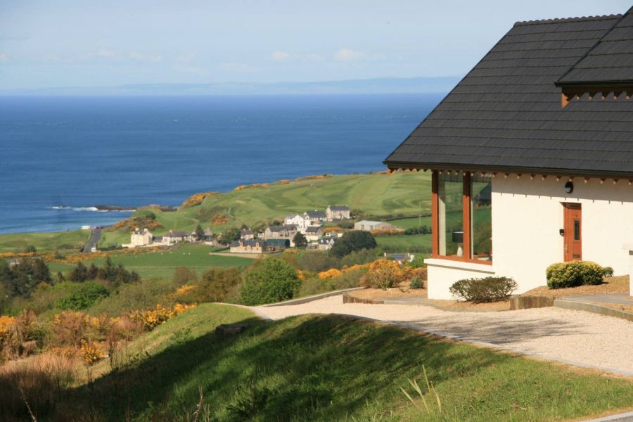 A scenic view from the Salthouse Hotel in County Antrim, Northern Ireland, overlooking rolling green hills, a coastal village, and the deep blue ocean. The modern hotel building with large windows and a sleek black roof is perched on a hillside, offering a tranquil and picturesque setting. The bright sunlight and clear skies enhance the beauty of the rugged coastline in the background.