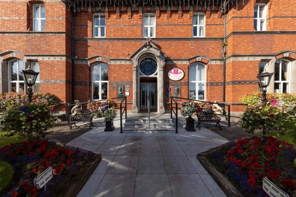 The charming red-brick facade of the Scholars Townhouse Hotel in Drogheda, Ireland, features elegant arched windows and a grand entrance with a decorative stone doorway. The entrance path is lined with vibrant flower arrangements, black wrought-iron benches, and classic lampposts, adding to the hotel's historic and welcoming atmosphere. Sunlight casts a warm glow on the building, highlighting its timeless architectural details.