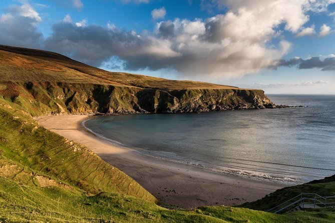 A breathtaking view of a secluded Irish coastline, featuring a crescent-shaped sandy beach nestled between rugged green cliffs. The calm waters of the Atlantic Ocean reflect the golden hues of the late afternoon sun, while dramatic clouds roll across the sky. A wooden staircase descends towards the shore, inviting visitors to explore the untouched beauty of this coastal paradise.