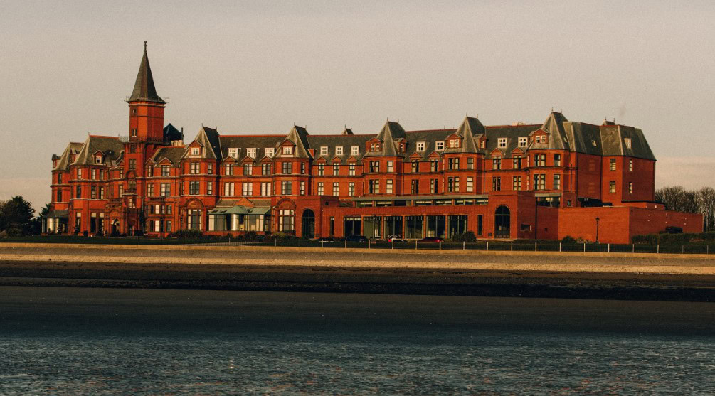 Exterior view of the Slieve Donard Hotel in Newcastle, Northern Ireland, showing the grand historic red-brick Victorian building with its clock tower and gabled roofs, overlooking the seafront under soft golden light; image credit to Booking.com.