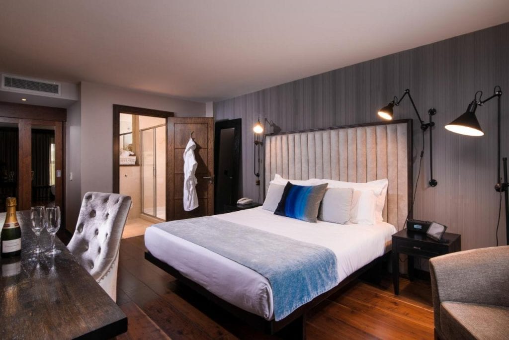 A stylish and modern hotel room at The Twelve Hotel in Galway, Ireland, featuring a plush king-size bed with a tufted headboard, soft blue and white bedding, and elegant black wall-mounted lamps. The room has a warm ambiance with dark wooden floors, a sleek work desk with a bottle of champagne and glasses, and an open doorway leading to a well-lit bathroom. A cozy and luxurious space designed for comfort and relaxation.