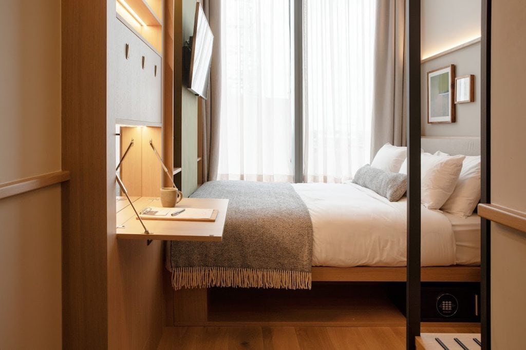 A modern, minimalist hotel room at the Wren Urban Nest in Dublin, Ireland, featuring a neatly made bed with white linens and a gray throw blanket, a wall-mounted TV, framed art, and a small fold-down desk with a mug and notebook. Photo courtesy of the Wren Urban Nest Hotel.