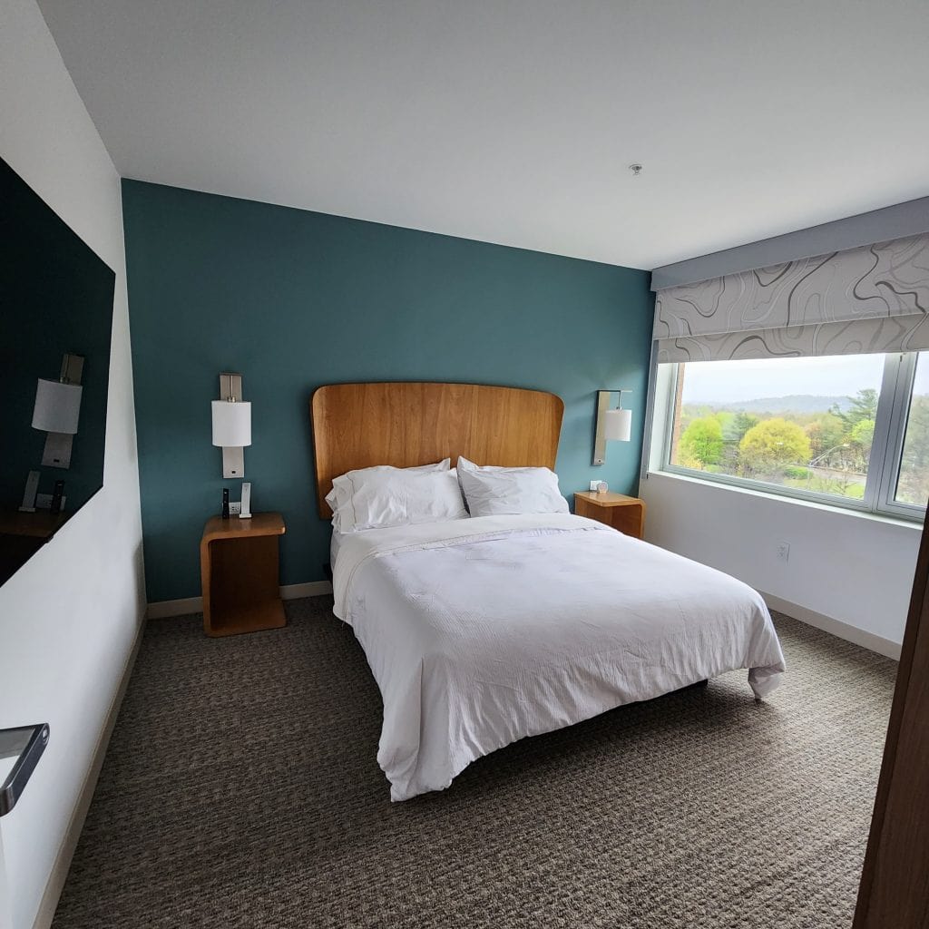 A modern bedroom at The Element Hotel in Asheville, NC, featuring a comfortable queen-sized bed with white linens and a wooden headboard against a teal accent wall. The room includes matching bedside tables with lamps and a large window offering natural light and views of greenery outside. The clean, minimalist decor creates a serene and inviting atmosphere.