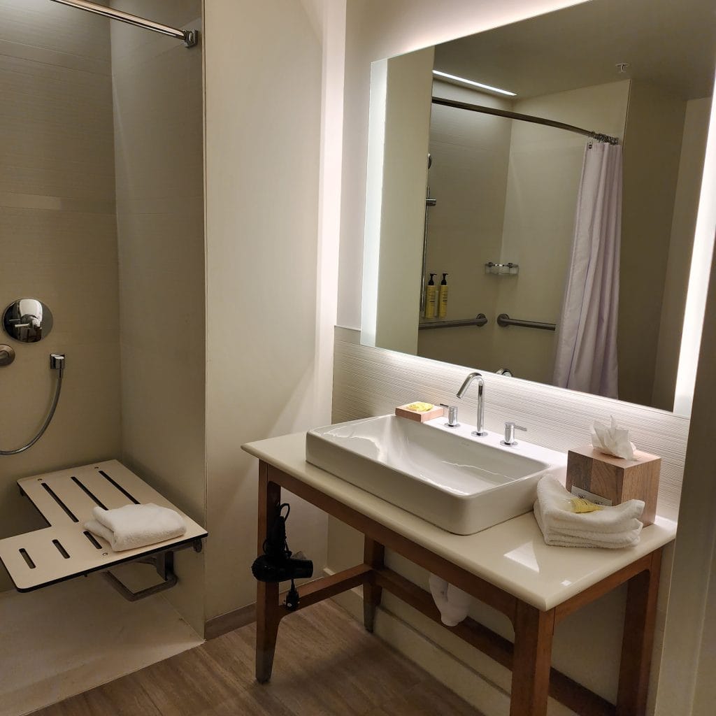 A sleek and accessible bathroom at The Element Hotel in Asheville, NC, featuring a walk-in shower with a fold-down bench and handheld showerhead. The vanity includes a modern rectangular sink, wooden accents, and a large backlit mirror. The clean design is complemented by thoughtful details such as rolled towels, toiletries, and a tissue box, ensuring comfort and functionality.
