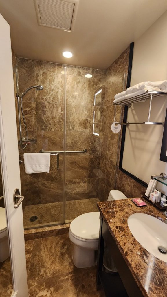 Bathroom in the Queen’s View Garden Room at The Royal Hawaiian Hotel in Waikīkī, featuring a walk-in shower with marble tile walls and glass doors, a modern toilet, a marble countertop with an under-mount sink, and a shelf with neatly stacked towels and amenities.