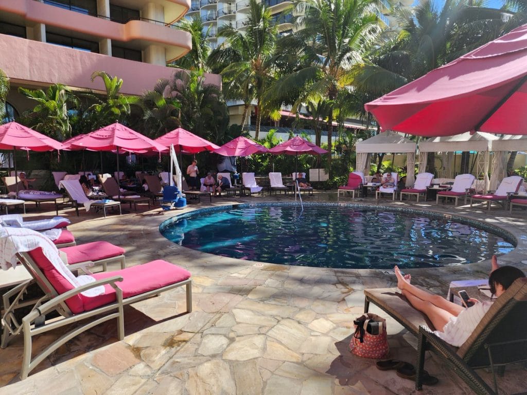 The Malulani Pool at The Royal Hawaiian Hotel in Waikīkī, Hawaii, surrounded by pink cushioned lounge chairs and bright pink umbrellas, with palm trees and cabanas adding tropical charm. Guests relax poolside in the sunshine near the hotel's signature pink architecture.
