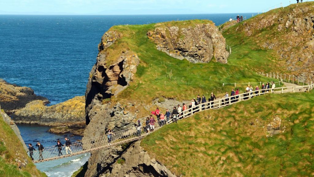 Tourists carefully walk across the narrow Carrick-a-Rede Rope Bridge, suspended high above the rocky cliffs and crashing waves below, connecting two grassy cliffs along the stunning North Antrim coast.