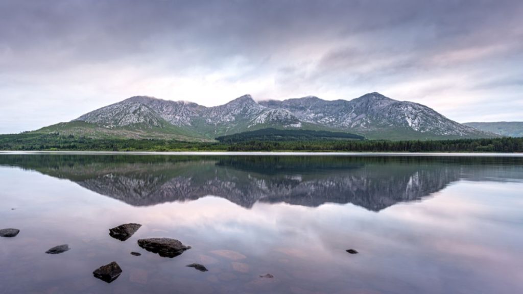 A serene view of Connemara National Park in Ireland, showcasing rugged mountains reflected in the still waters of a lake. The sky is overcast with soft, moody clouds, complementing the lush greenery and rocky peaks. A few scattered stones peek out from the water in the foreground, adding depth to the peaceful landscape.