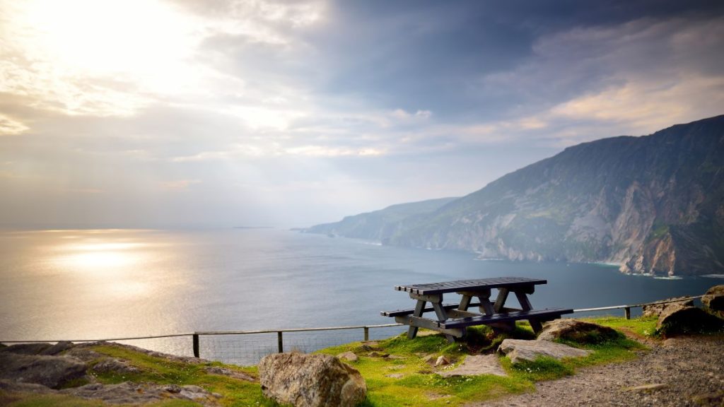 A serene coastal view in Donegal, Ireland, featuring a wooden picnic table on a grassy cliffside overlooking the Atlantic Ocean. Sunlight filters through a cloudy sky, casting a golden glow over the water, with towering cliffs stretching into the distance. A breathtaking spot for reflection and scenic beauty.