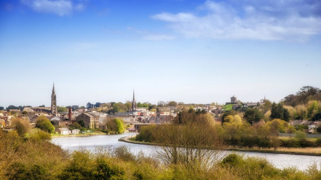 A panoramic view of Drogheda, Ireland, taken by Anthony Murphy via MythicalIreland.com, showing the River Boyne winding through the town with church spires, historic buildings, and lush green trees under a bright blue sky.