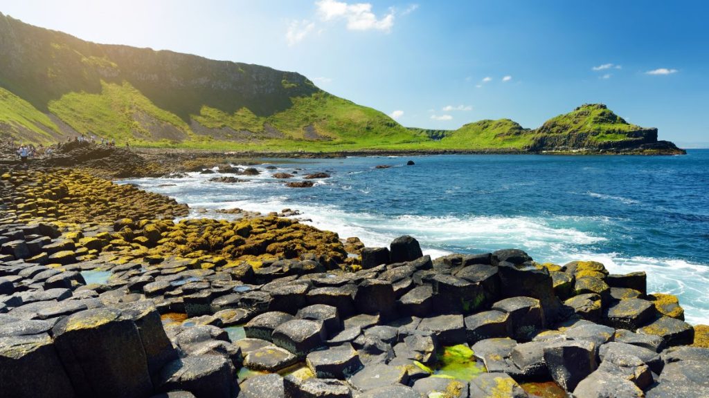 A rugged coastal landscape near the Giant’s Causeway in County Antrim, Northern Ireland, features black volcanic rocks along the shoreline with waves crashing against them. In the background, a green cliffside rises dramatically, partially illuminated by sunlight. The deep blue sky and scattered white clouds enhance the breathtaking and untamed beauty of this iconic location.
