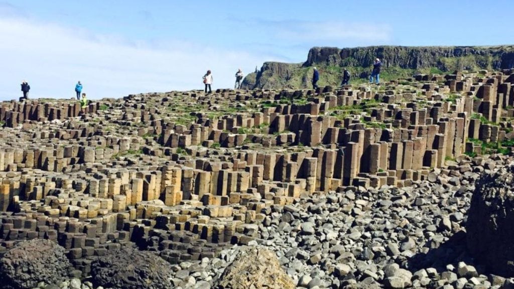 Tourists walking atop the unique interlocking basalt columns of the Giant’s Causeway in Northern Ireland under a clear blue sky.