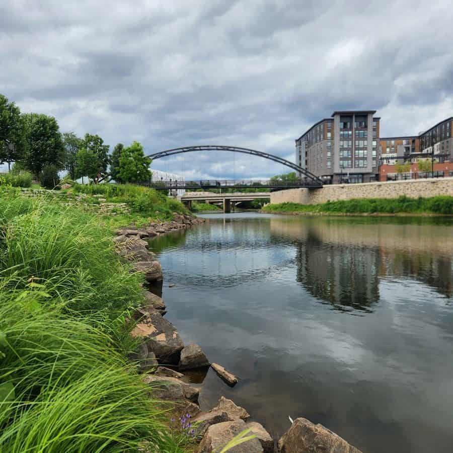 Scenic view of the River Lights Bridge arching over the Chippewa River at Phoenix Park in Eau Claire, Wisconsin. Modern apartment buildings rise in the background while lush green grass, trees, and rocky riverbanks frame the calm, reflective water under a cloudy sky.