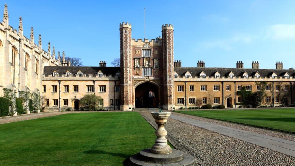 Spacious courtyard at Trinity College in Dublin, featuring a manicured green lawn, a central stone ornament, and a grand arched building with decorative spires and symmetrical windows under a clear blue sky.