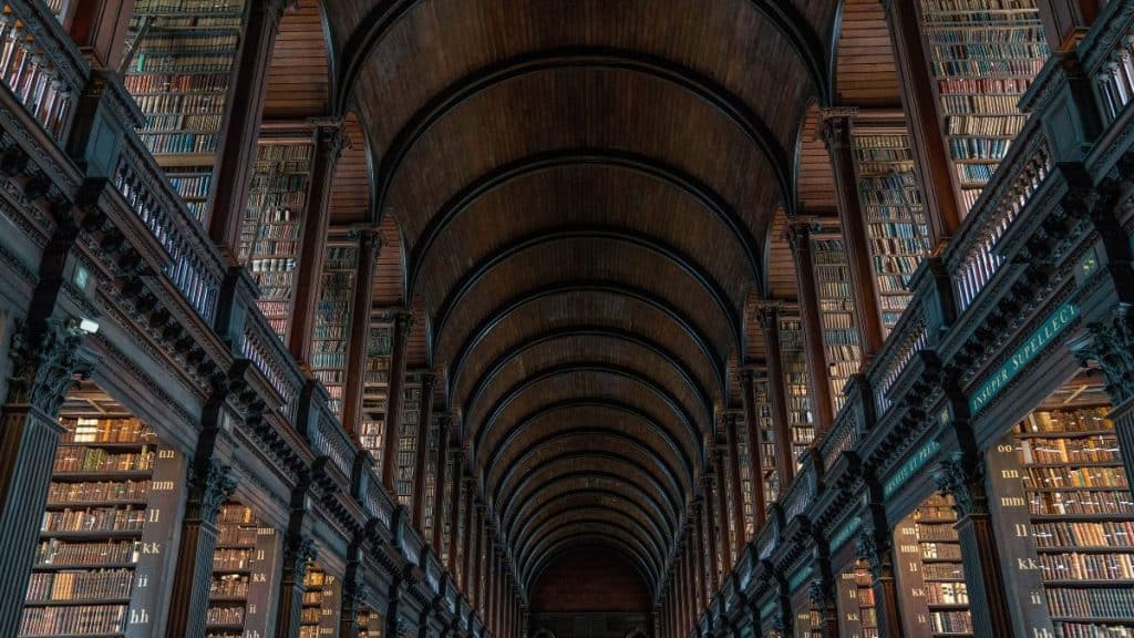 The iconic Long Room inside Trinity College Library in Dublin, Ireland, featuring a vaulted wooden ceiling, rows of tall bookshelves filled with ancient texts, and a dim, atmospheric lighting.