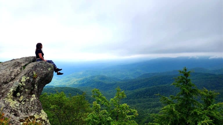 A woman sitting on Blowing Rock overlooking the mountains and forest below.