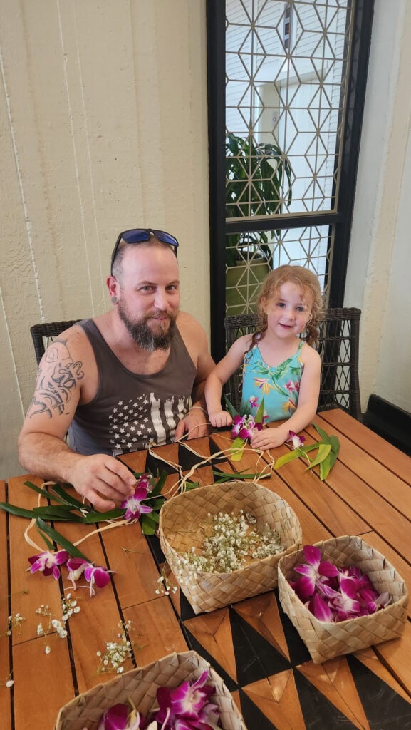 A father and daughter smile while making leis at a wooden table covered with purple orchids, baby's breath, and greenery at the Royal Hawaiian Hotel. Woven baskets hold loose flowers, and both are engaged in the hands-on cultural activity.







