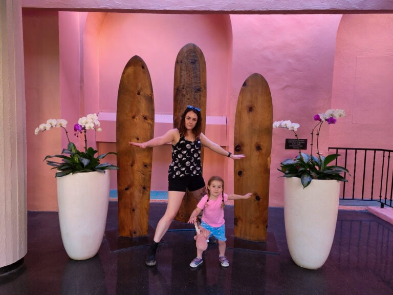 A mother and daughter strike playful surfing poses in front of three wooden surfboards displayed against a vibrant pink wall at the Royal Hawaiian Hotel. They stand between large white planters filled with blooming orchids, capturing a fun and tropical family moment.