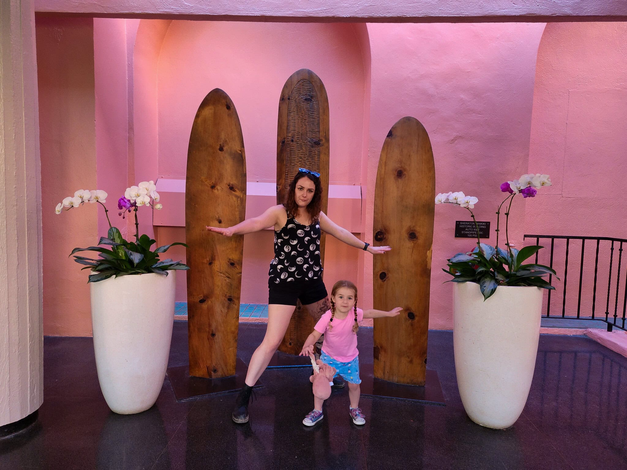 A mother and daughter strike playful surfing poses in front of three wooden surfboards displayed against a vibrant pink wall at the Royal Hawaiian Hotel. They stand between large white planters filled with blooming orchids, capturing a fun and tropical family moment.