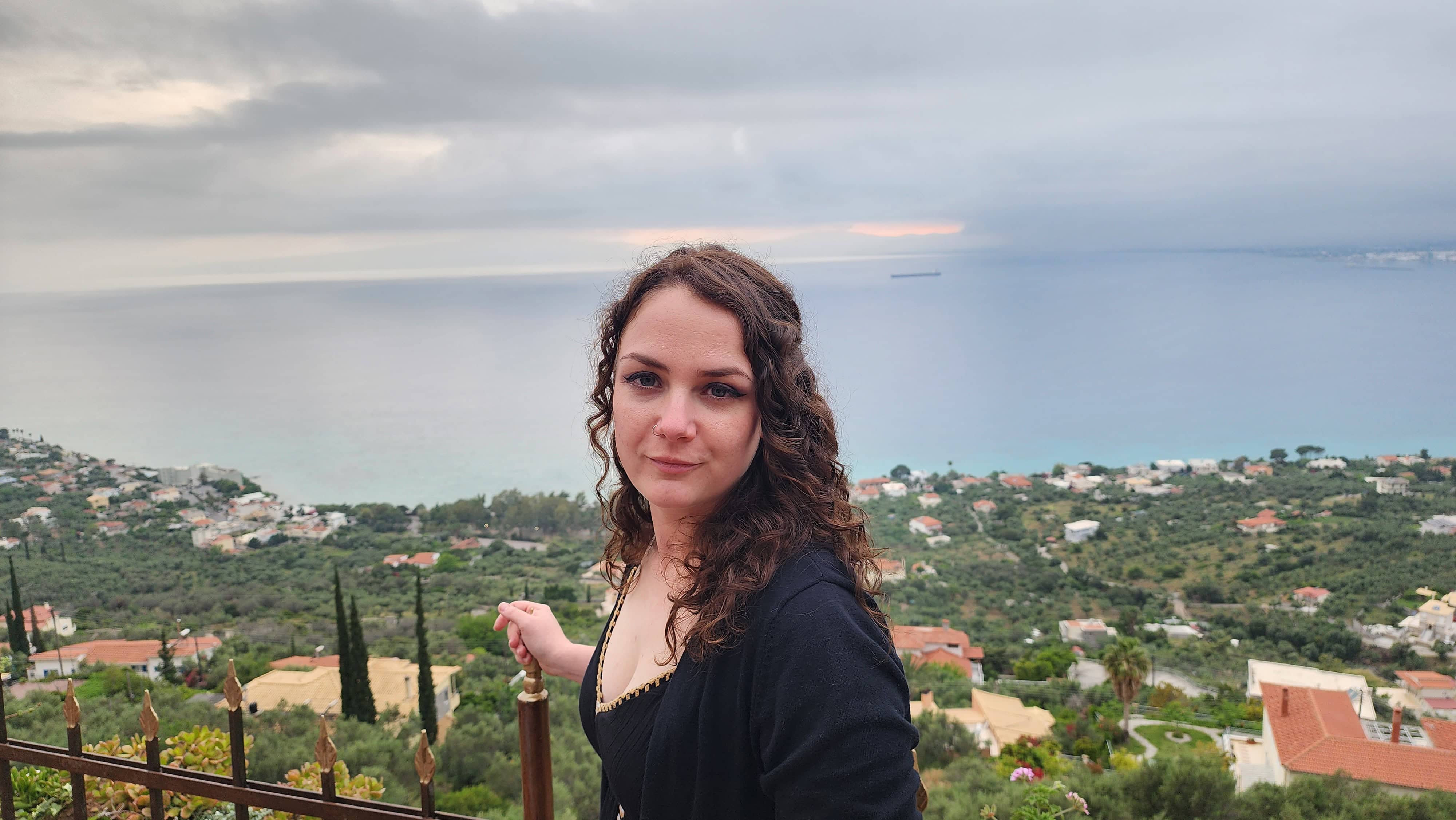 Portrait of a woman standing at the top of a cliffside in Kalamata, Greece, overlooking a scenic view of the sea, distant coastal homes, and lush greenery under a cloudy sky. Phoenyx posing for a picture at the edge of a cliffside in Kalamata, Greece.