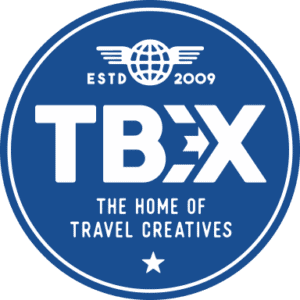 Circular blue logo for TBEX featuring bold white text that reads “TBEX” with an arrow incorporated into the letter X. Above it is a globe with wings and the text “ESTD 2009,” and below it reads “The Home of Travel Creatives” with a small star at the bottom.