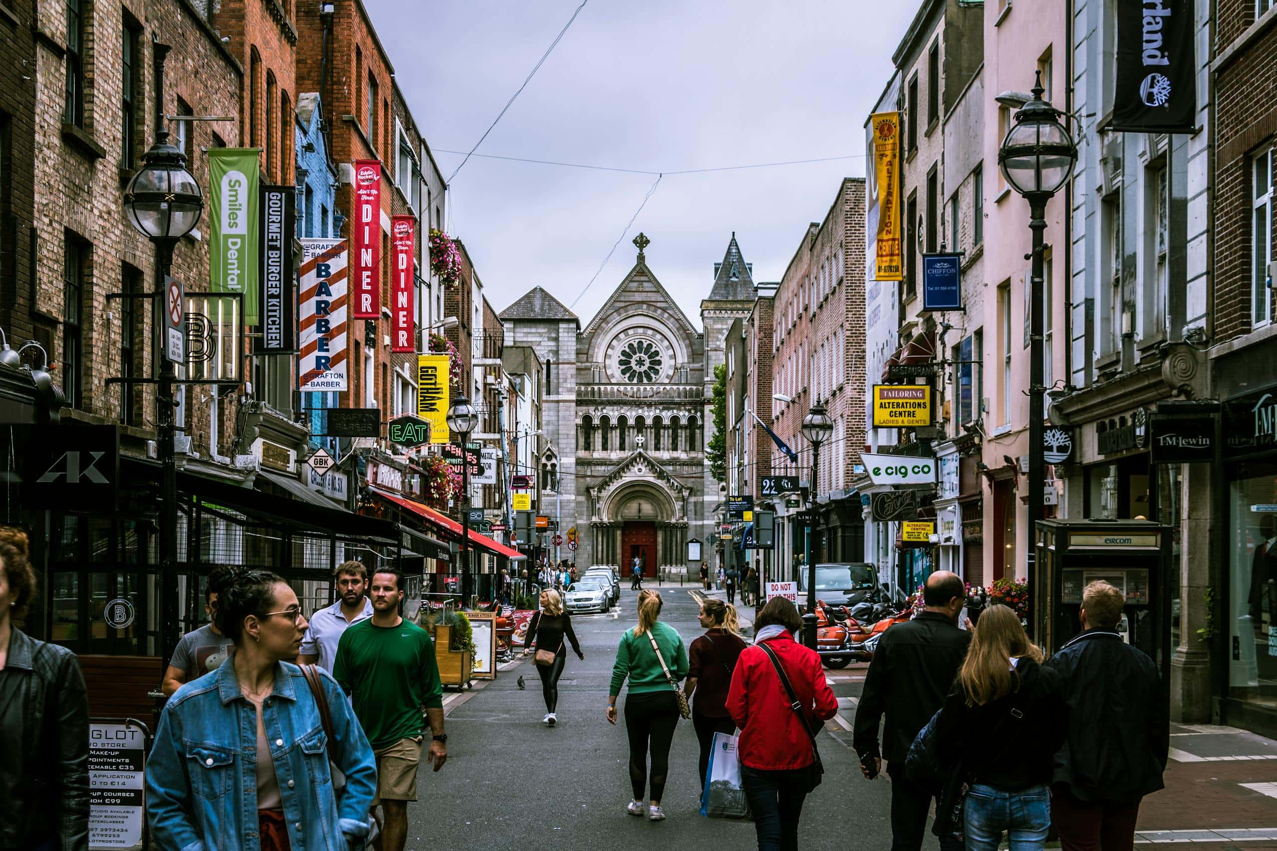 A lively street in Dublin, Ireland, photographed by Lukas Kloeppel via Pexels, showing pedestrians walking between historic brick buildings lined with colorful shop signs, with a grand stone church featuring a rose window at the end of the street.