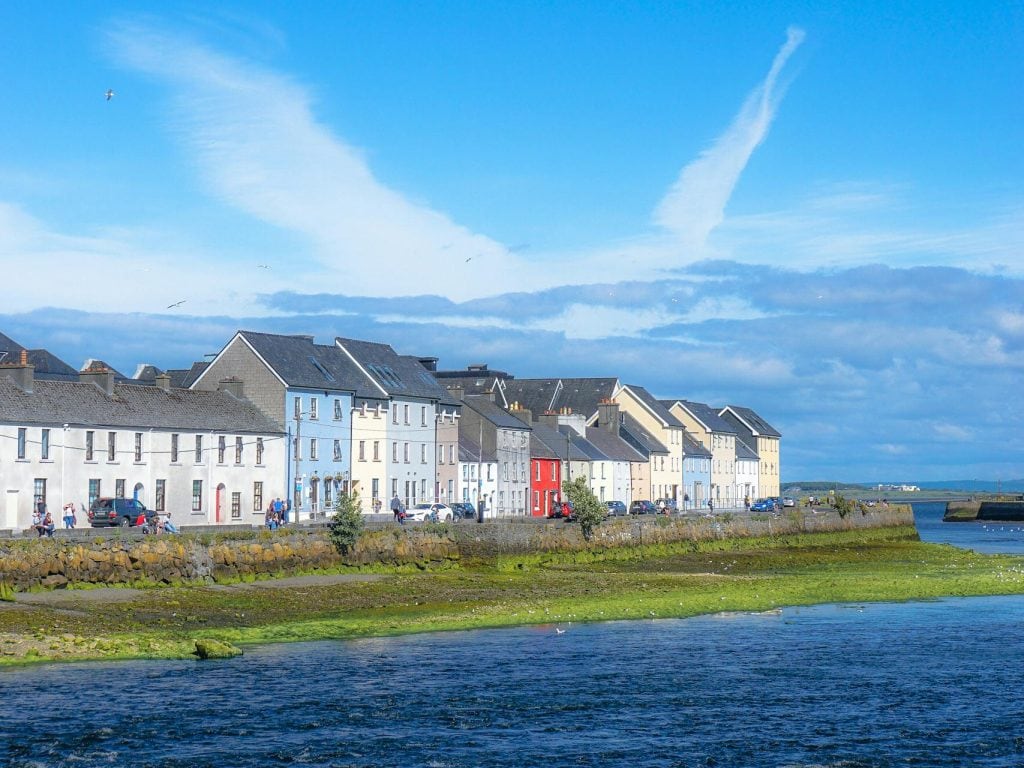 A scenic view of the colorful houses along the waterfront in Galway, Ireland, with a bright blue sky and wispy white clouds overhead. The vibrant row of homes contrasts with the deep blue water, while people stroll along the stone wall and seagulls fly above. The scene captures the charm of Galway’s coastal beauty and lively atmosphere.