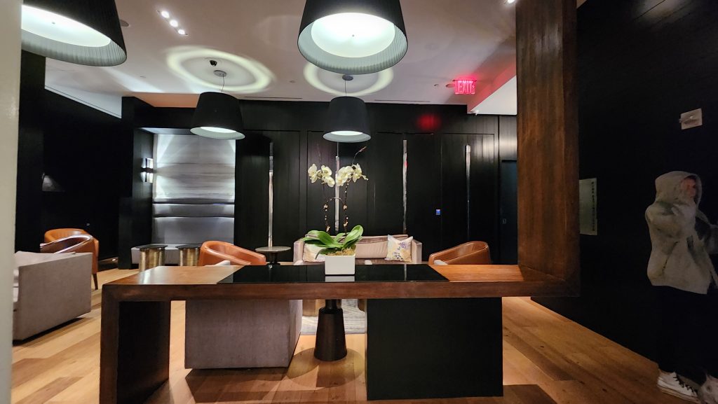 Stylish and modern lobby of the Andaz 5th Avenue in NYC, featuring warm wood accents, contemporary black pendant lighting, sleek furniture, and a central table adorned with an elegant orchid arrangement.
