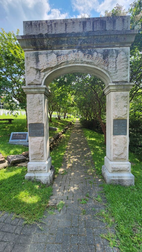 Stone memorial archway at Oakwood Cemetery in Raleigh, NC, with engraved text and plaques on either side, leading to a shaded brick path surrounded by green grass and trees under a bright, sunny sky.