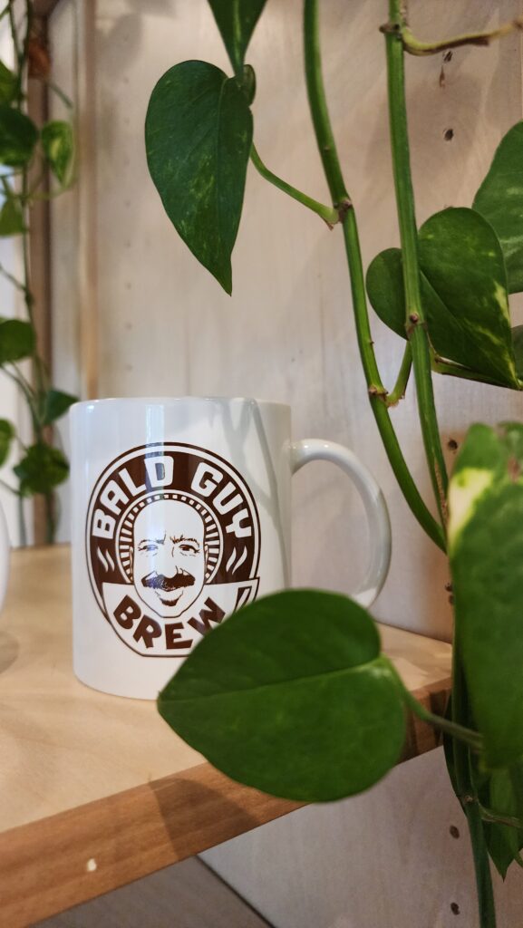 A white coffee mug with the Bald Guy Brew logo, featuring a smiling bald man with a mustache, placed on a wooden shelf surrounded by green leafy plants at Bald Guy Brew in Blowing Rock.