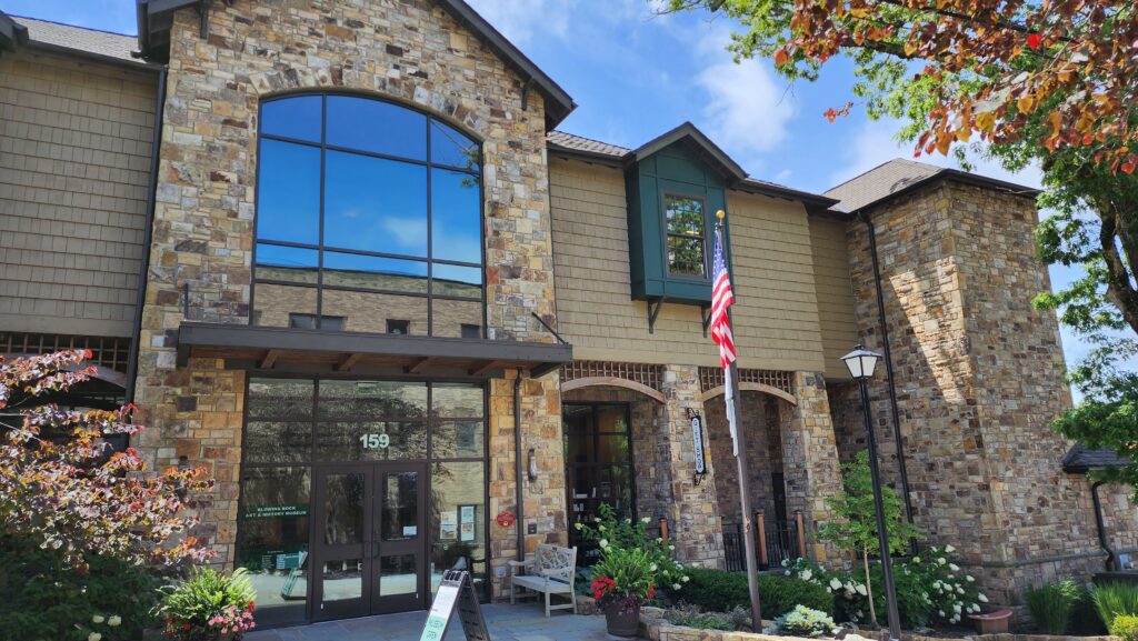 The Blowing Rock Art & History Museum, a stone and wood-paneled building with large glass windows reflecting the blue sky, surrounded by vibrant flowers and greenery. An American flag stands proudly at the entrance alongside a welcoming bench.
