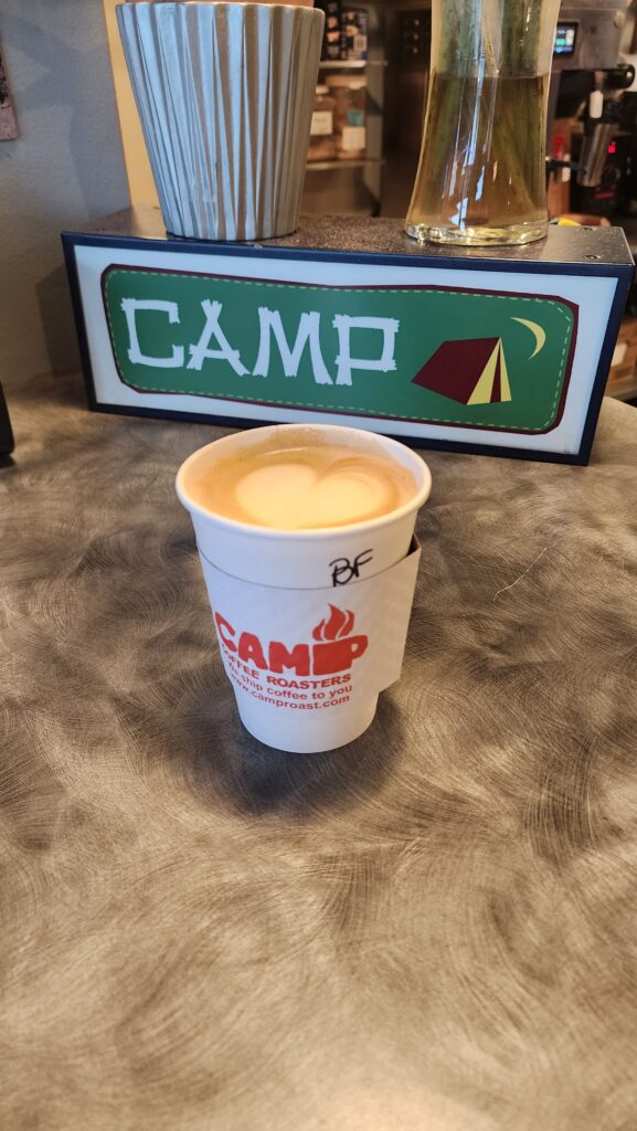 A cheerio latte served in a Camp Coffee Roasters branded cup, placed on a metallic countertop with a decorative 'CAMP' sign featuring a tent and crescent moon in the background.