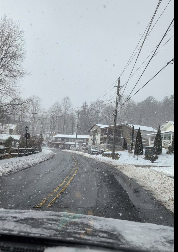 A snowy street scene in downtown Blowing Rock, NC, captured from a car, with snow-covered buildings, trees, and sidewalks lining the winding road as snowflakes fall gently.