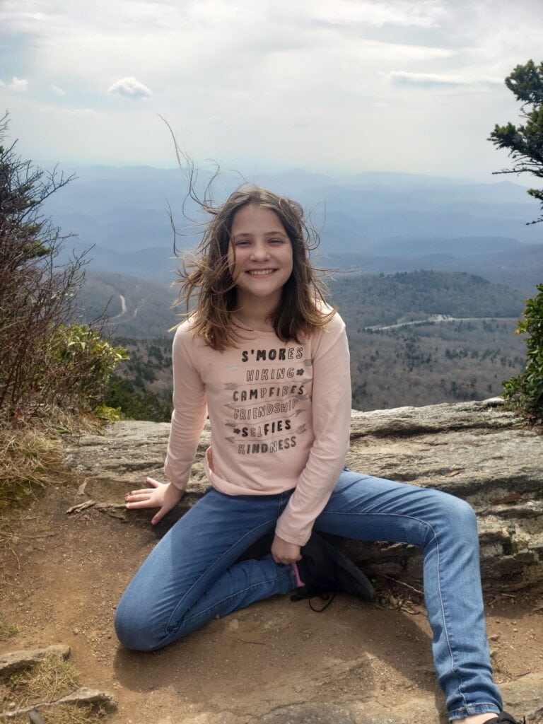 Evelyn smiles while sitting on a rocky ledge at Grandfather Mountain, North Carolina, with wind blowing her hair. The scenic background features layered Blue Ridge Mountain ridges stretching into the distance under a partly cloudy sky, creating a picturesque outdoor moment.