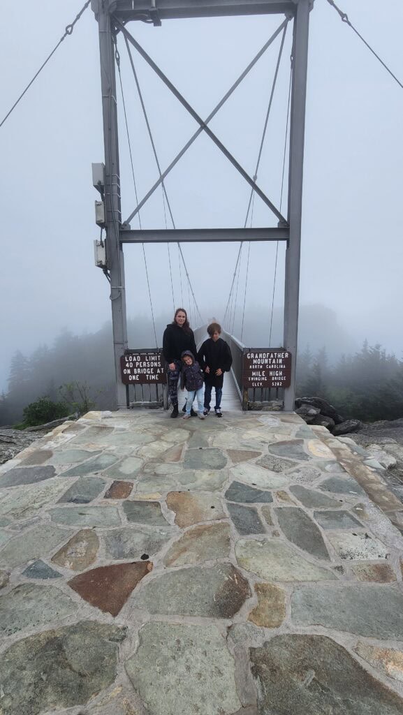 A family stands at the start of the Mile High Swinging Bridge at Grandfather Mountain, North Carolina, surrounded by dense fog. The stone pathway leading to the bridge and the steel bridge frame are visible, with misty trees in the background, creating a dramatic and atmospheric scene.