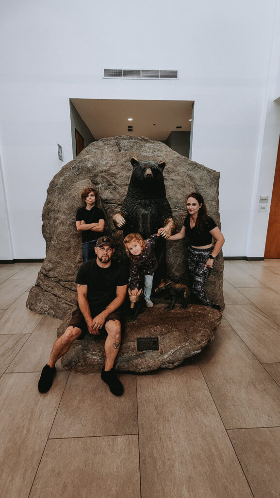 Phoenyx and her family pose with a life-sized bear statue at the Wilson Center for Nature Discovery at Grandfather Mountain, North Carolina. The family is gathered around the detailed statue set against a large stone background, inside a bright and modern space with tiled floors and high ceilings.