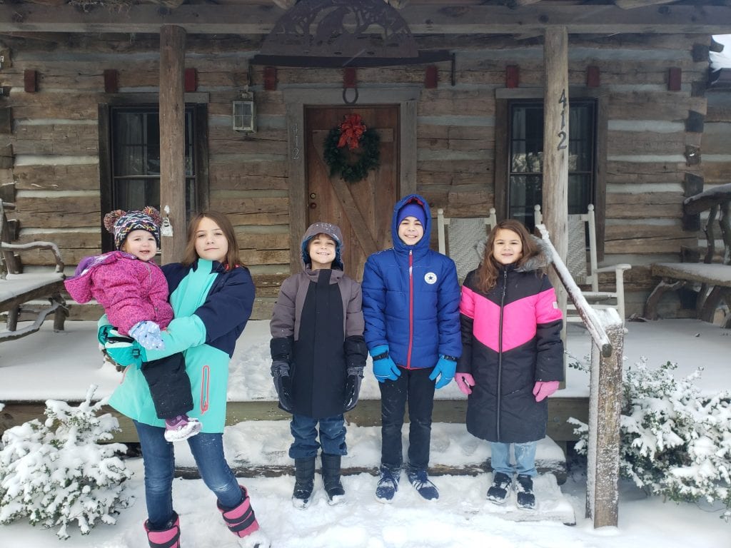 Five children from a blended family standing together in front of a snow-covered log cabin during a Christmas vacation in North Carolina, dressed in colorful winter coats, with festive decorations on the cabin door.