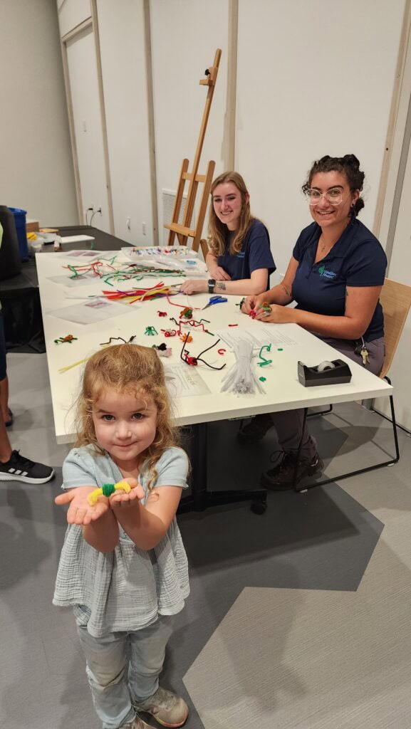 River proudly shows off a colorful craft she created during Animal Enrichment Day at Grandfather Mountain Nature Park. Behind her, two staff members sit at a table covered with crafting supplies, smiling as they assist other visitors in making enrichment items.