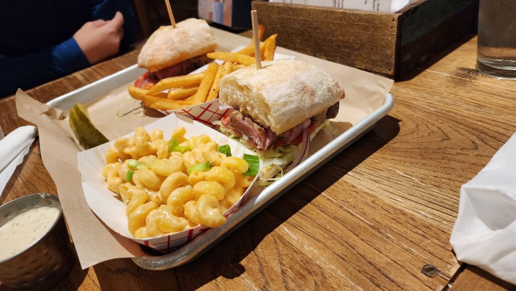 A lunch tray at Hellbender Bed and Beverage featuring a steak sandwich with lettuce, tomato, and onion on ciabatta bread, accompanied by crispy French fries, creamy mac and cheese topped with green onions, and a pickle spear
