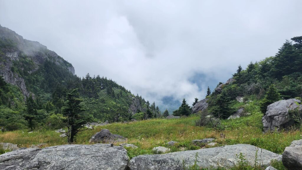 A breathtaking view from Grandfather Mountain, featuring a lush green meadow framed by rocky outcrops and evergreen trees. Mist and low-hanging clouds drift between the surrounding cliffs and valleys, creating a serene and mystical atmosphere.