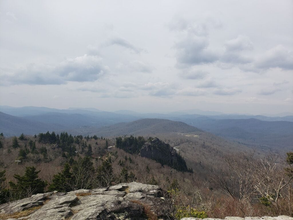 A scenic view of the rolling Blue Ridge Mountains taken from the Mile High Swinging Bridge at Grandfather Mountain, North Carolina. The landscape features rugged rock formations in the foreground, sparse trees transitioning to dense forests, and layers of misty mountain ridges under a cloudy sky.
