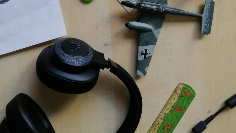 A pair of noise-canceling headphones for airplanes resting on a wooden surface, alongside a small model airplane with camouflage paint and a ruler featuring frog designs. The scene includes casual items, emphasizing travel or hobby themes.