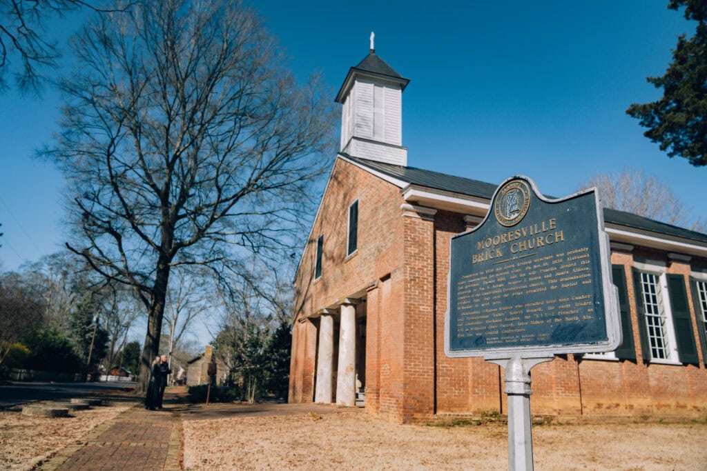 Historic marker in front of the Mooresville Brick Church in Mooresville, Alabama, with the red-brick structure and white steeple visible under a clear blue sky. A person stands in the background on the brick path, reading another sign near the large bare trees lining the quiet street.