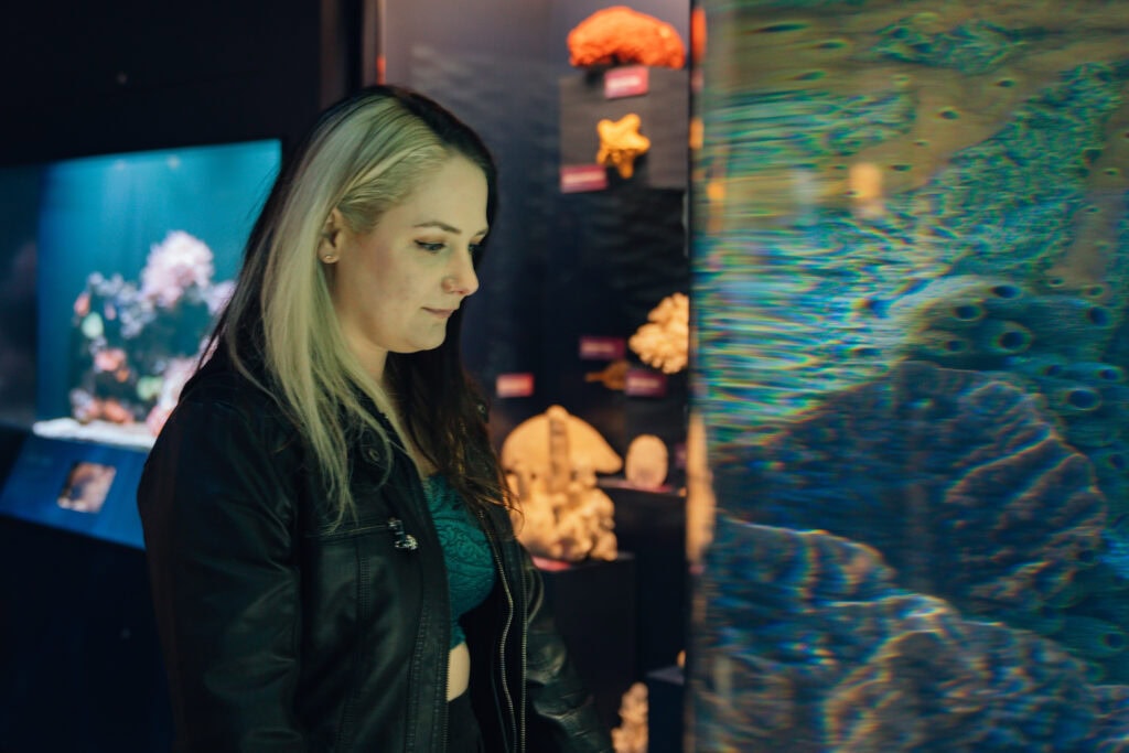 Phoenyx stands near a glowing aquarium tank at the Cook Museum of Natural Science in Decatur, Alabama, surrounded by colorful coral displays and underwater life exhibits.