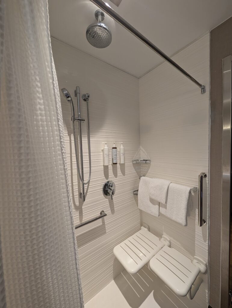 Clean, accessible shower at AC Hotel Downtown Huntsville featuring a fold-down seat, dual shower heads, wall-mounted toiletries, and handrails.