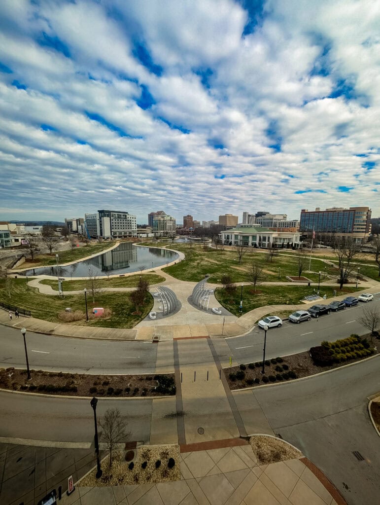 A wide-angle daytime view of Big Spring International Park in downtown Huntsville, Alabama, featuring the amphitheater-style seating, reflective pond, walking paths, and city skyline under a dramatic, cloud-filled sky.